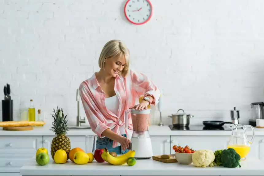 happy young woman preparing tasty nutritious smoothie in blender