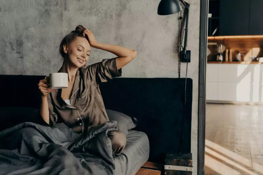 Portrait of young relaxed woman in stylish home suit waking up with cup of coffee in bed