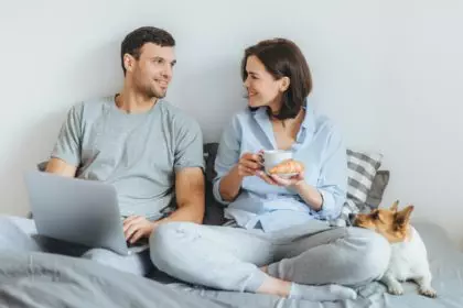 Couple sit together on bed, use laptop computer, have pleasant conversation between each other