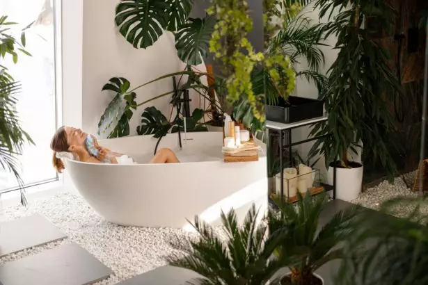 Relaxed young woman bathing in modern bathroom interior decorated with tropical plants